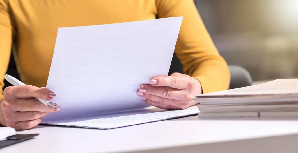 Des mains tiennent un stylo au-dessus d'un document imprimé, avec une tasse de café et des papiers éparpillés sur le bureau
